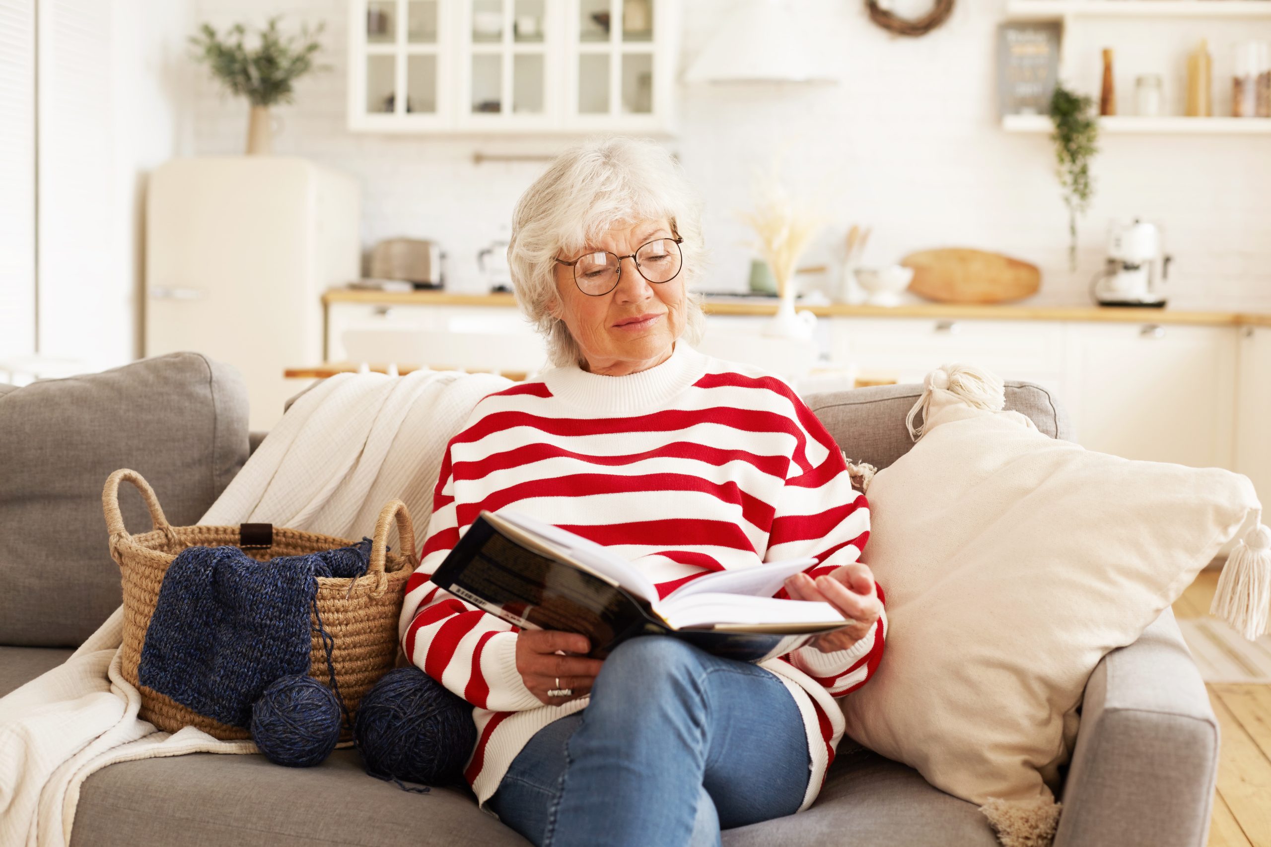 Portrait of stylish attractive female pensioner in round eyeglasses relaxing at home with good book. Joyful elderly woman wearing spectacles, reading bestseller, sitting comfortably on sofa